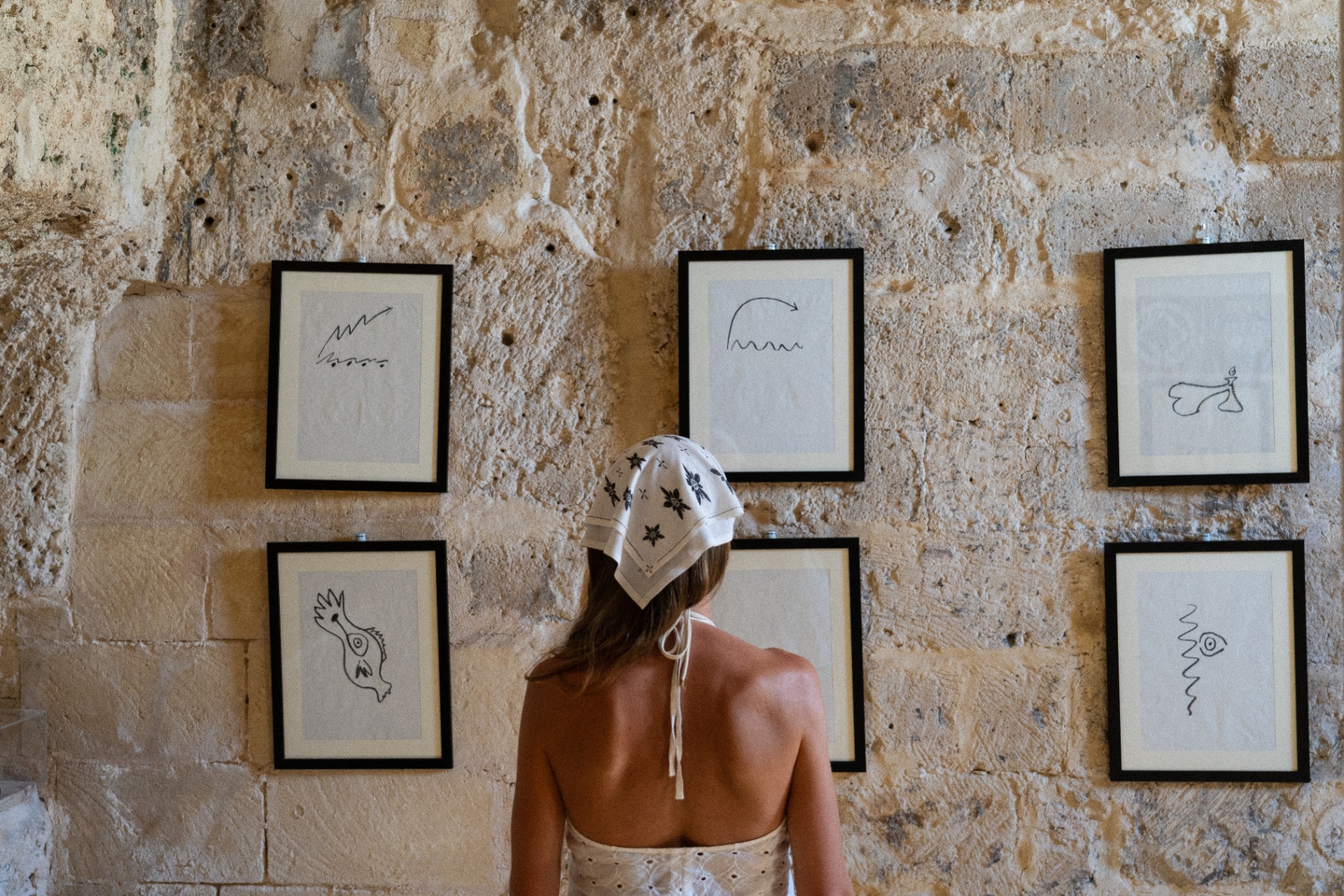 Une femme de dos face à un mur de pierre orné de dessins encadrés